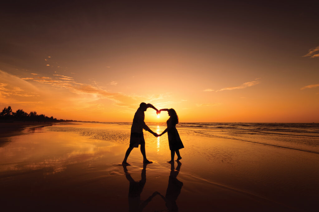 Silhouette of couple making heart shape with arms on beach at su